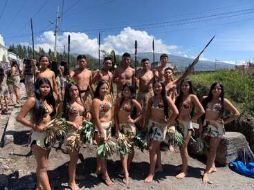 Um grupo de jovens, homens e mulheres, veste trajes tradicionais com elementos decorativos, segurando lanças e folhagens, posando ao ar livre sob um céu ensolarado, com montanhas ao fundo.