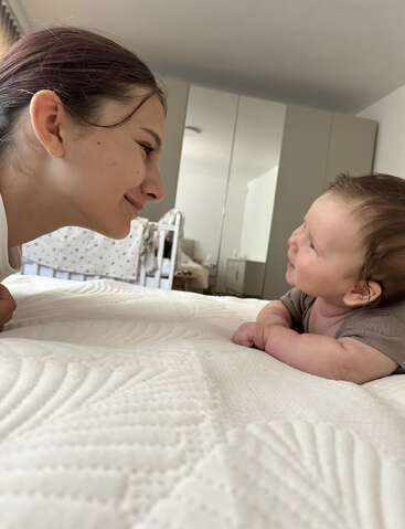 A woman and a baby lie facing each other on a bed, smiling lovingly. The background shows a crib and mirrored wardrobe, suggesting a cozy, intimate setting.