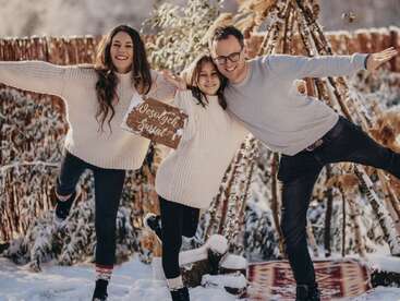 A happy family poses playfully in the snow, wearing cozy sweaters. They hold a sign reading “Wesołych Świąt” (Merry Christmas) and smile warmly outdoors.