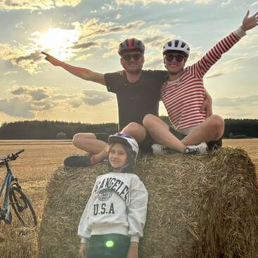 Three people wearing helmets enjoy a sunny countryside scene, sitting on a hay bale, smiling, with bicycles nearby, arms outstretched, radiating happiness and togetherness.