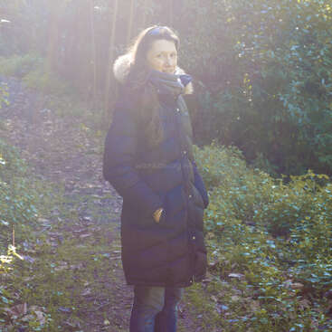 A woman stands on a forest path, wearing a long dark winter coat and scarf. Sunlight filters through trees, creating a serene and peaceful atmosphere.