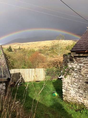 L'image représente un paysage serein avec un arc-en-ciel au-dessus d'un champ herbeux, avec un mur de pierre, une clôture en bois et des collines lointaines sous un ciel gris.