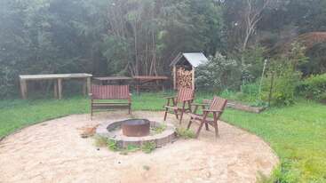 This image shows a cozy outdoor fire pit surrounded by wooden chairs, a bench, lush greenery, a woodpile, a table, and peaceful natural surroundings.