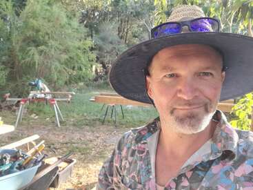 A man wearing a wide-brim hat and sunglasses smiles outdoors. Behind him, woodworking tools and lumber indicate a construction or DIY project amidst green trees.
