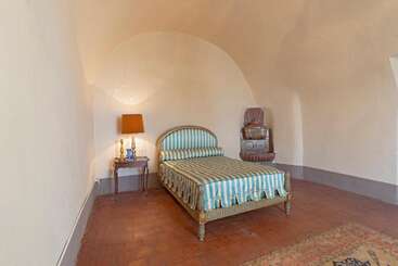 A minimalist bedroom features a bed with blue and gold striped bedding, a small side table with a lamp, a chair, and terracotta-tiled floors.