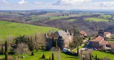 A picturesque stone chateau with turrets sits amidst rolling green hills and scattered trees, surrounded by other rustic buildings under a partly cloudy, bright sky.
