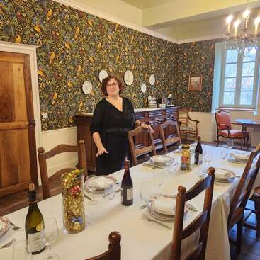 A woman stands in a warmly decorated dining room with vibrant floral wallpaper. The elegant table is set with wine, plates, glasses, and festive centerpieces.