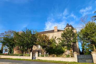 This image shows a charming stone building with a turret, surrounded by trees and greenery, under a bright blue sky with wispy clouds, behind a gate.