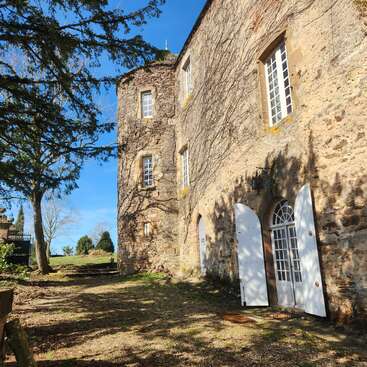 A rustic stone building with arched doors and white shutters, surrounded by trees and sunshine, exudes a peaceful, historic charm amidst a natural setting.