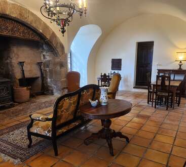 This cozy, historic room features a large stone fireplace, vintage furniture, a round wooden table, chandelier, tiled floor, and soft lighting giving a warm, inviting atmosphere.