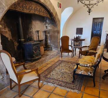 This cozy room features a large stone fireplace, wood stove, ornate chairs, a patterned rug, and vintage furniture, evoking a warm, rustic, historical ambiance.