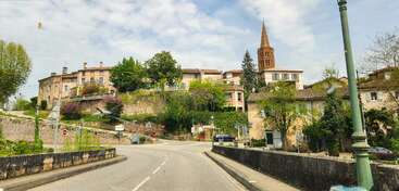 This image shows a quaint European village with stone buildings, a church with a tall steeple, greenery, winding road, and street signs under a partly cloudy sky.