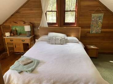 A cozy bedroom with wooden walls, a neatly made bed with white linens, bedside tables, a vintage vanity, folded towels, bright windows, and decorative wall art.