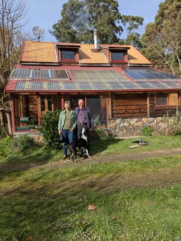 Um casal está com seu cachorro preto e branco na frente de uma casa rústica de madeira com painéis solares e chaminé, cercada por vegetação e árvores.