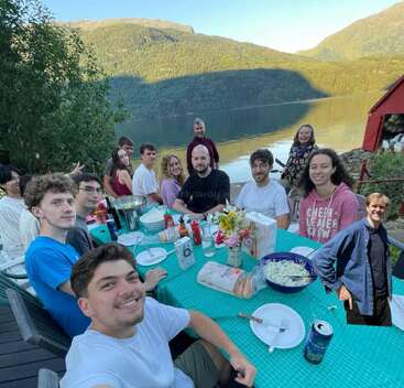 Un groupe d'amis se réunit autour d'une table en plein air, savourant un repas au bord d'un magnifique lac, entourés par les montagnes et la nature, souriants et heureux ensemble.
