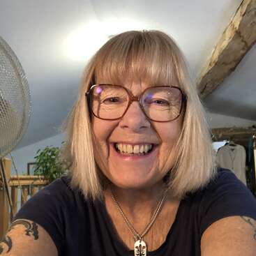 L'image montre une femme aux cheveux blonds et aux lunettes, portant un t-shirt noir et un collier, prenant un selfie dans une pièce où se trouvent un ventilateur et des plantes.