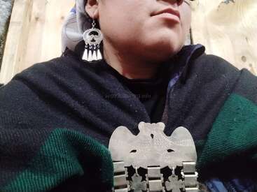 A person wears traditional clothing, including a dark shawl, silver earrings, and a decorative silver brooch. The background is wooden, suggesting an indoor rustic setting.