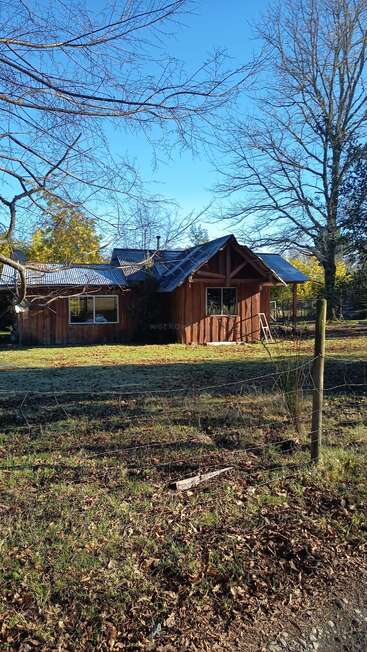 A rustic wooden cabin sits quietly in a grassy yard, surrounded by bare trees and a wire fence, under a clear blue sky. Peaceful countryside scene.