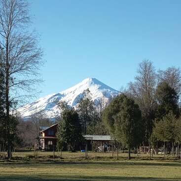 A peaceful rural landscape features a snow-capped mountain in the background, surrounded by trees. In the foreground, there is a cozy wooden house and open field.