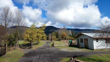 Uma paisagem rural apresenta uma estrada de cascalho, uma casa branca, cercas de madeira, árvores vibrantes, grama verde, montanhas distantes e um céu azul brilhante com nuvens fofas.