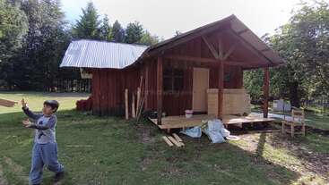 A young boy plays near a rustic wooden cabin surrounded by trees. Construction materials are scattered around, and sunlight filters gently through the outdoor scene.