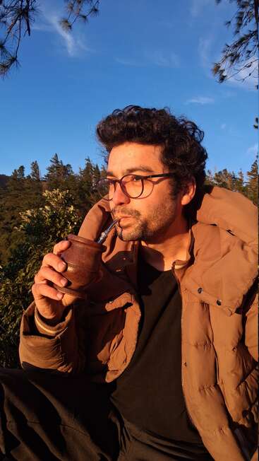 Un hombre con gafas y chaqueta marrón está sentado al aire libre, bebiendo mate de una calabaza. La luz del sol ilumina su rostro. Al fondo, pinos y cielo azul.