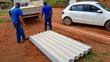 Deux hommes en chemise bleue déchargent des matériaux de construction, notamment des tôles ondulées et des poutres en bois, d'un camion situé à côté d'une voiture blanche sur une surface en terre battue.