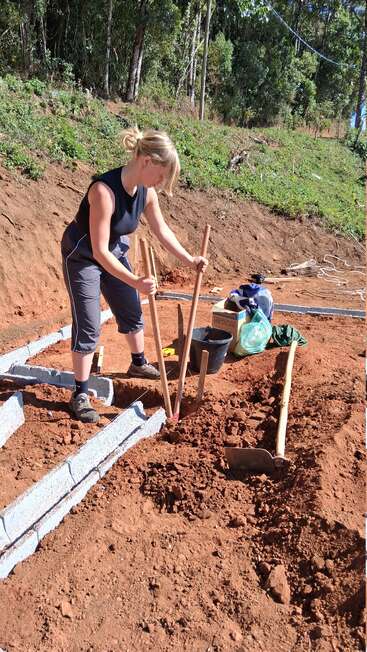 Une femme travaille en plein air sur un chantier de construction, creusant dans la terre rouge à l'aide de poteaux en bois. Des outils, des blocs et des sacs sont éparpillés à proximité, sous la lumière du soleil.