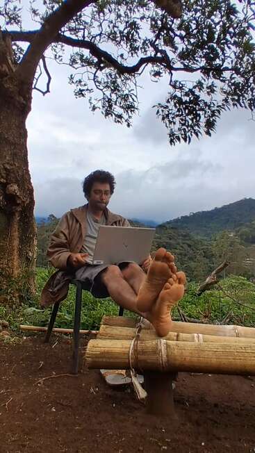 Un homme est assis en plein air sous un grand arbre, travaillant sur un ordinateur portable. Ses pieds nus reposent sur un banc en bambou, avec des collines verdoyantes derrière lui.