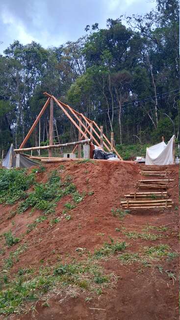 Une maison en bois est en cours de construction sur un terrain surélevé, avec des marches en bambou. Une forêt dense et un ciel nuageux sont à l'arrière-plan.