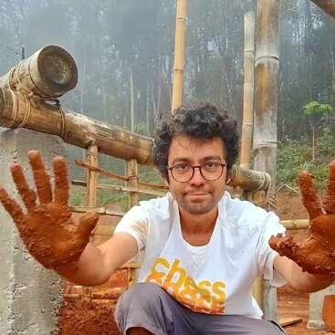 Un homme aux lunettes et aux cheveux bouclés, vêtu d'un t-shirt blanc, montre ses mains couvertes de boue alors qu'il travaille sur une construction en bambou et en ciment à l'extérieur.