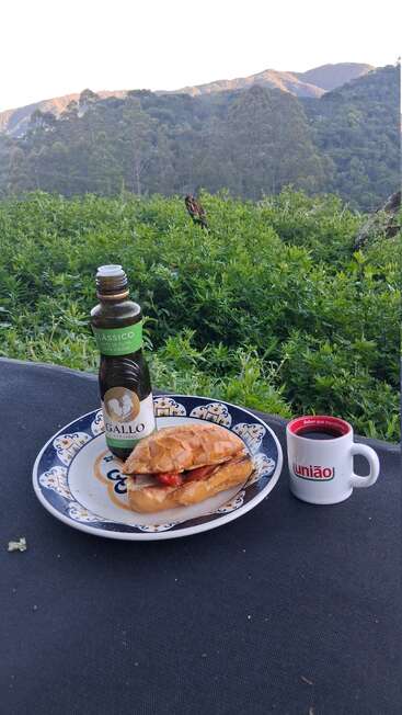A scenic outdoor breakfast setup with a sandwich, olive oil bottle, and coffee cup on a plate, surrounded by lush greenery and mountains in the background.