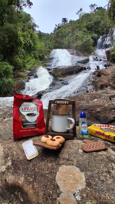 A picturesque waterfall serves as backdrop to a cozy picnic setup featuring coffee, cookies, biscuits, and sugar, creating a serene and inviting outdoor moment.