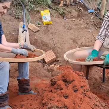 Zwei Leute sitzen draußen und sieben rötliche Erde durch große runde Siebe. Mit Handschuhen und Freizeitkleidung arbeiten sie zusammen, umgeben von Erde, Werkzeugen und einem Erdhaufen.