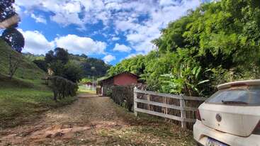 Une scène champêtre rustique mettant en scène un chemin de terre, une voiture blanche garée, des arbres verts, une clôture en bois et une petite maison rouge sous un ciel d'un bleu éclatant.