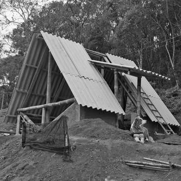 En una colina de tierra, rodeado de árboles, hay un refugio rústico con estructura en forma de A sin terminar. Un perro yace cerca. Unos escalones de madera descienden por la ladera de tierra.