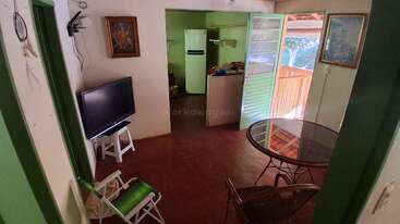 Ce salon confortable est équipé d'une télévision, de chaises pliantes, d'une table à manger ronde en verre, d'une petite cuisine, de deux tableaux et d'une porte ouverte donnant sur une véranda en bois.