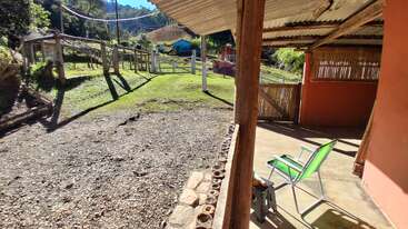 Une véranda rustique, où trône une chaise verte, donne sur un jardin clôturé, une pelouse et des collines boisées baignées par un soleil radieux, créant ainsi une atmosphère champêtre paisible.