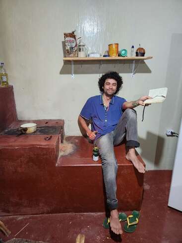 A man with curly hair sits barefoot on a raised platform in a rustic kitchen, smiling, holding a notebook and a drink, with sandals on the floor.