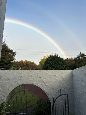 Un arc-en-ciel brillant traverse un ciel clair au-dessus d'un mur blanc texturé et d'un portail en fer forgé, entouré d'arbres verts et d'un sentier de jardin paisible.