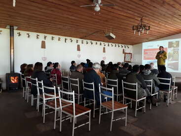 Un grupo de personas está sentado en el interior, escuchando con atención a un ponente que habla junto a una pantalla de proyección. La sala tiene techos de madera y una estufa de leña.