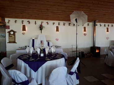 La imagen muestra un salón de banquetes decorado con mesas redondas, manteles blancos y morados, una elegante vajilla, una mesa con la tarta, una estufa de leña y un paraguas fotográfico.
