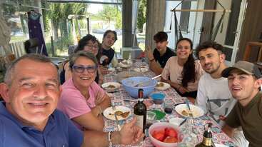 Eine Gruppe von acht Leuten, die lächeln und gemeinsam an einem bunten, gemusterten Tisch drinnen essen. Die Stimmung ist freundlich, entspannt und voller Freude.