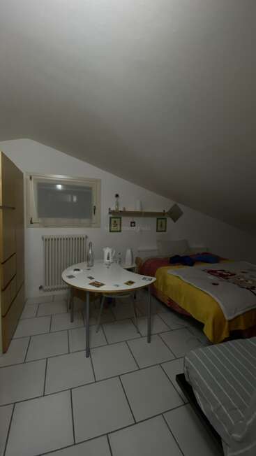 Das Bild zeigt ein gemütliches Dachzimmer mit schräger Decke, einem Bett, einem kleinen Tisch mit Stühlen, einer Kommode, Fliesenboden und schlichter Einrichtung.