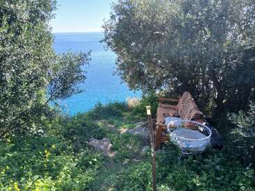 Un coin salon extérieur confortable avec un canapé et une table nichés parmi les arbres verts, surplombant une mer bleue magnifique sous un ciel clair, invitant à la détente.