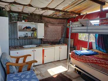 This cozy room features a small kitchenette, bunk beds, a patterned armchair, colorful textiles, open shelves, and abundant natural light streaming through large windows.
