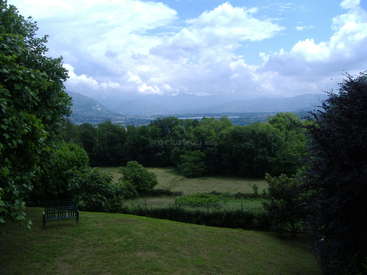 A imagem retrata uma paisagem serena com um campo verde exuberante, árvores e um banco, tendo como pano de fundo montanhas distantes sob um céu nublado.