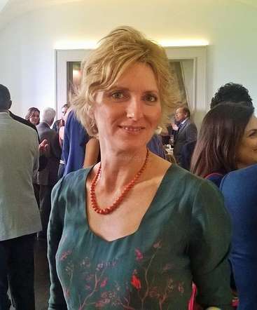 La imagen muestra a una mujer de pelo corto y rubio, con un top verde y un collar rojo, de pie en una sala llena de gente, probablemente en un acto social.