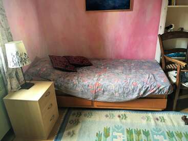 A cozy bedroom features a single bed with floral bedding, two patterned pillows, a wooden nightstand, a lamp, a chair, and a pastel pink wall.