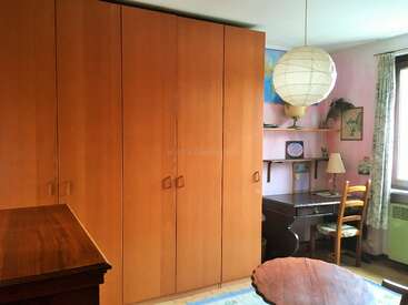 This cozy room features large wooden wardrobes, a dark ergonomic desk, a wooden chair, a round table, paper lantern light, map, and sunny window.